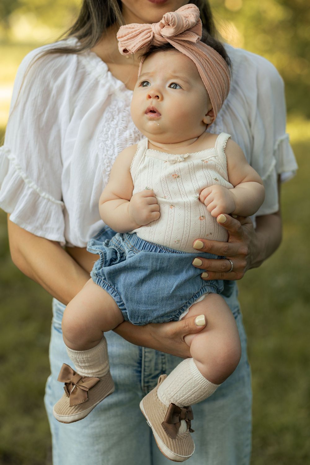 Mother holding baby girl in pink bow headband, white top, denim shorts, and bow shoes outdoors.