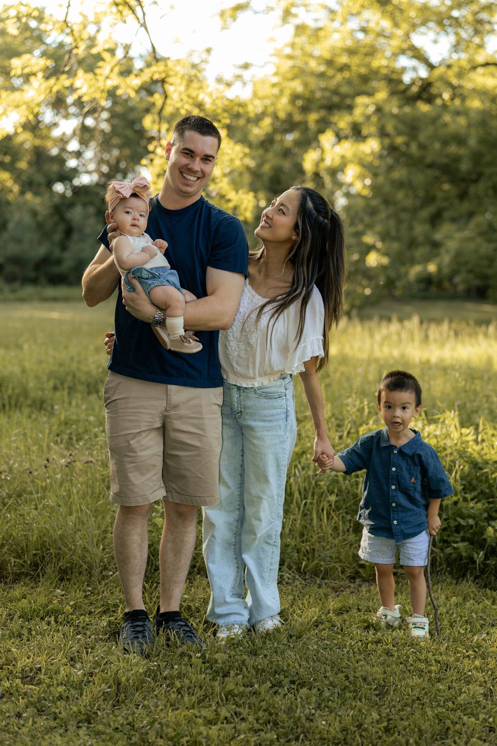 Happy family of four posing outdoors in a sunny green park, parents holding young children, smiling together.