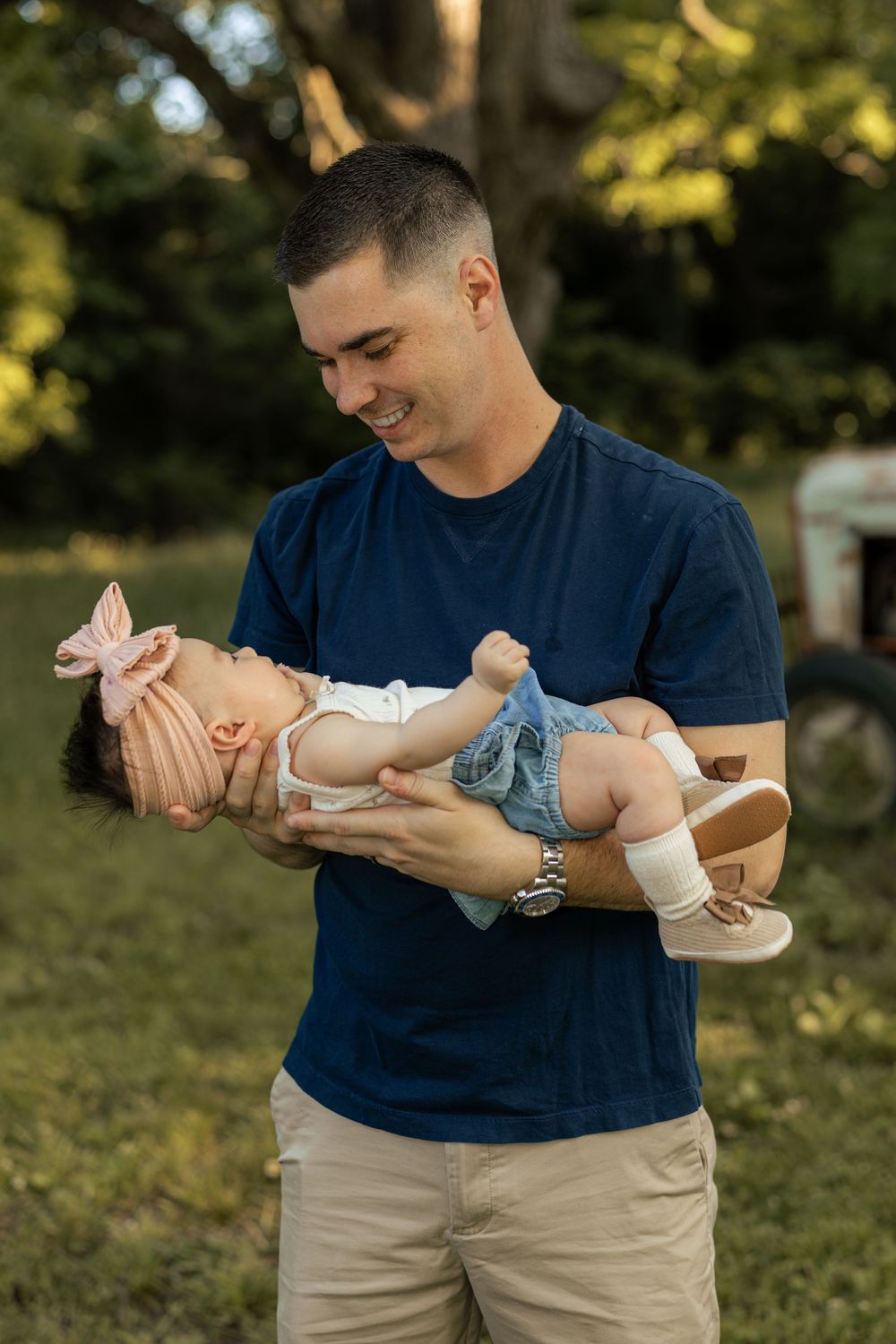 A smiling man in a navy shirt holds a baby girl in denim overalls and pink bow outdoors in golden light.
