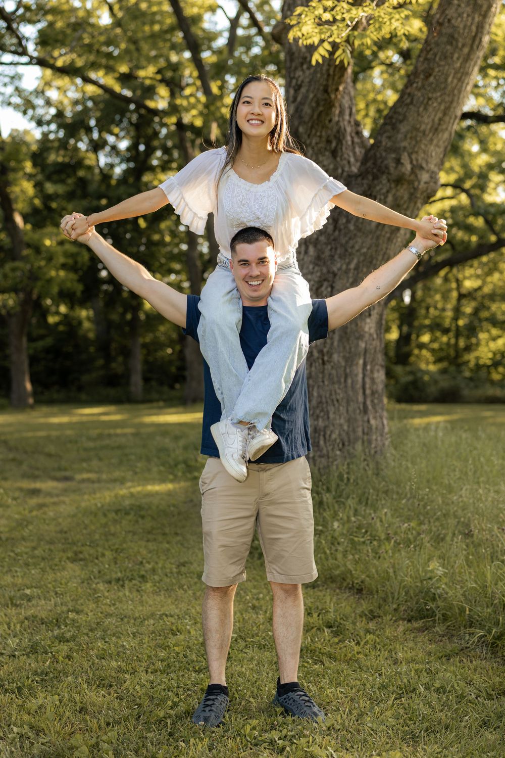 Happy couple in a park, man carrying woman on his shoulders, both smiling outdoors among green trees.