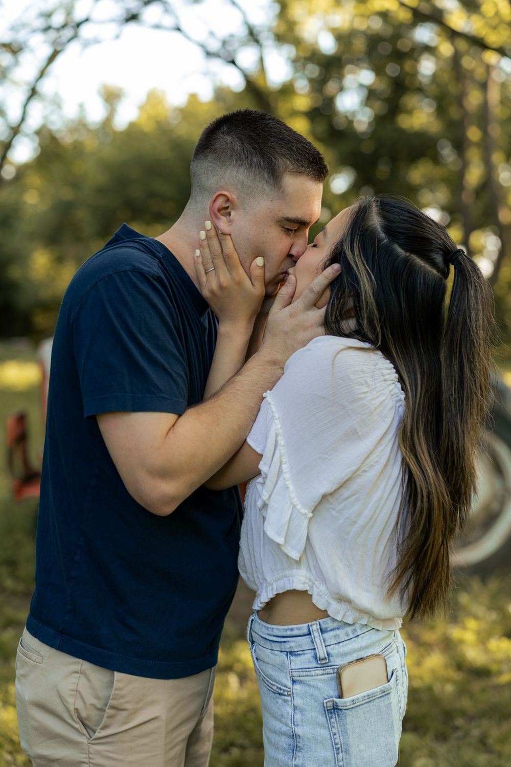 Couple sharing a romantic kiss outdoors in a park, man in navy shirt and woman in white top and jeans.
