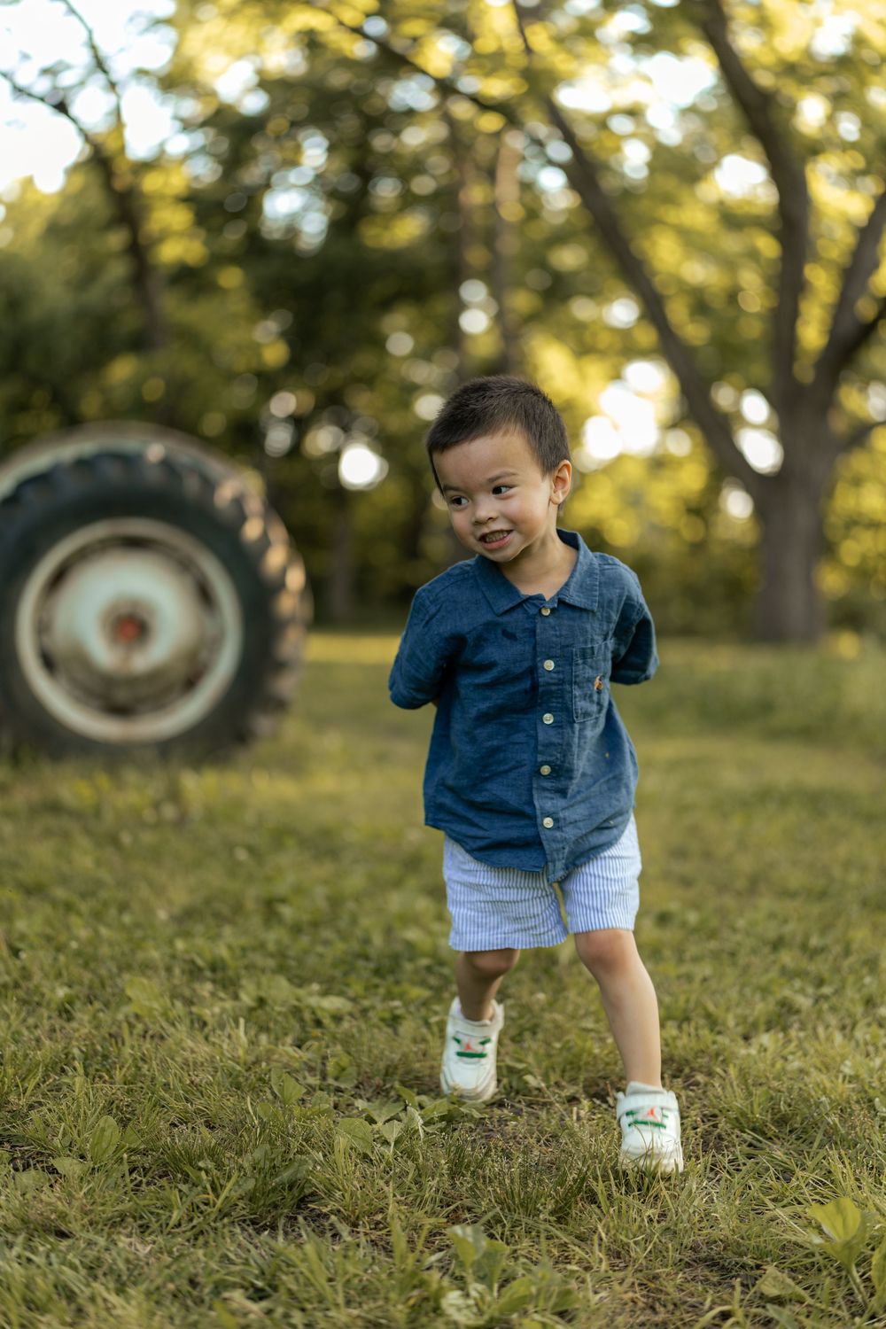 A toddler boy in a denim shirt and white shorts walks on grass near a tractor in a sunlit outdoor setting.