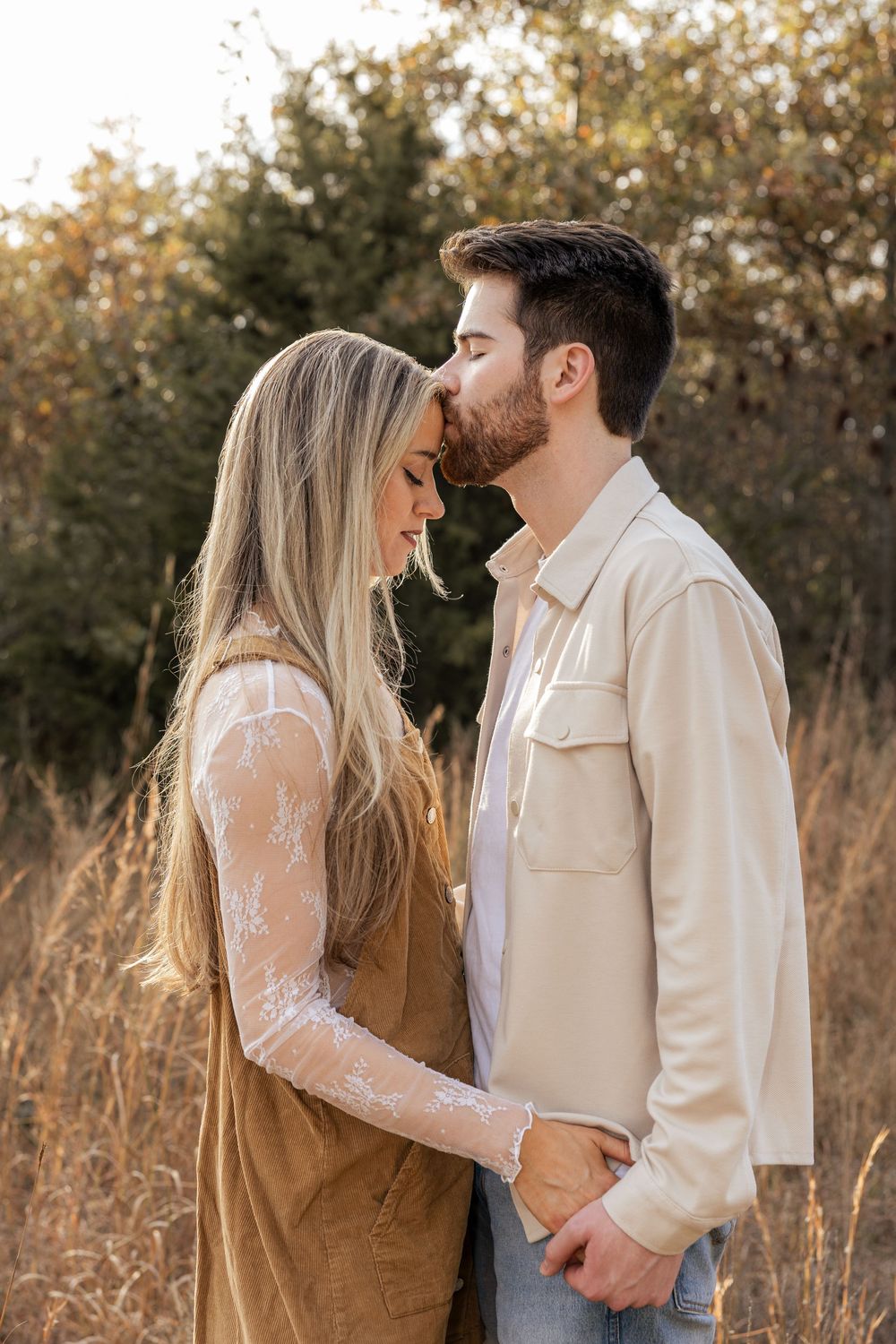 A couple shares a tender forehead kiss in a golden field, the woman in lace and suede, the man in a beige shirt.
