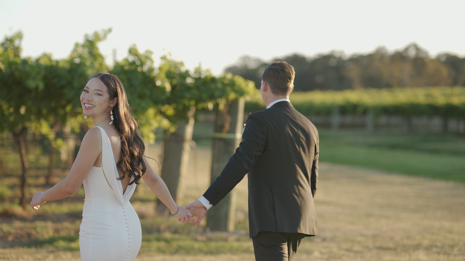 Wedding couple walking hand in hand through a scenic vineyard at sunset.