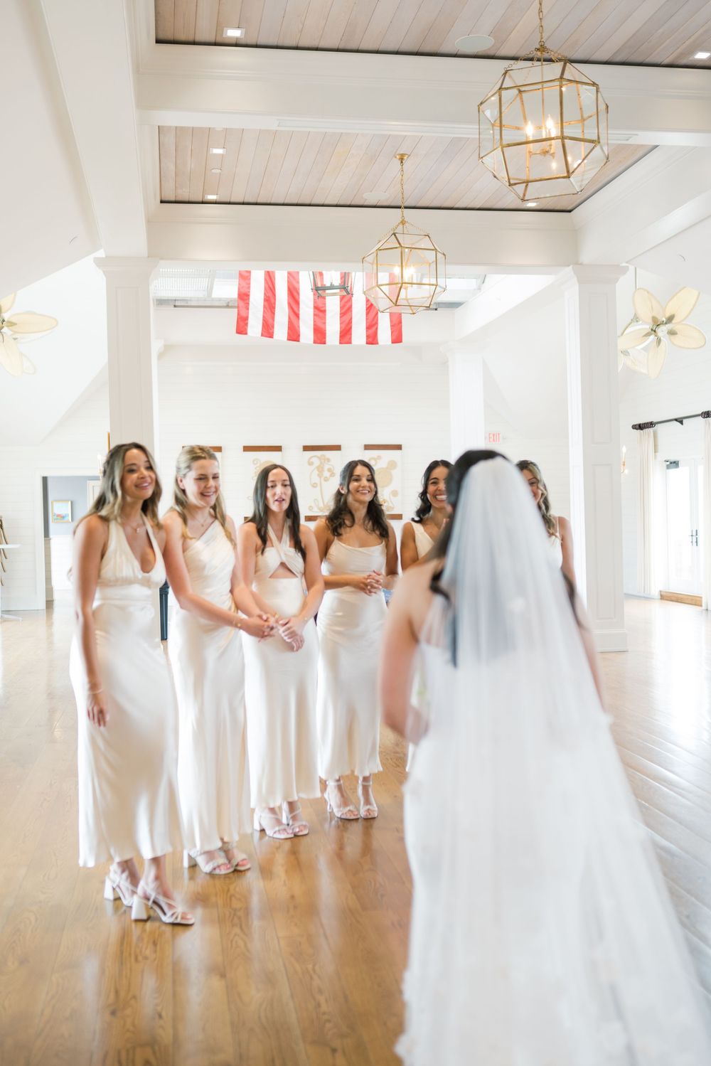 Wedding party in white dresses gathered in bright room with hanging lanterns.