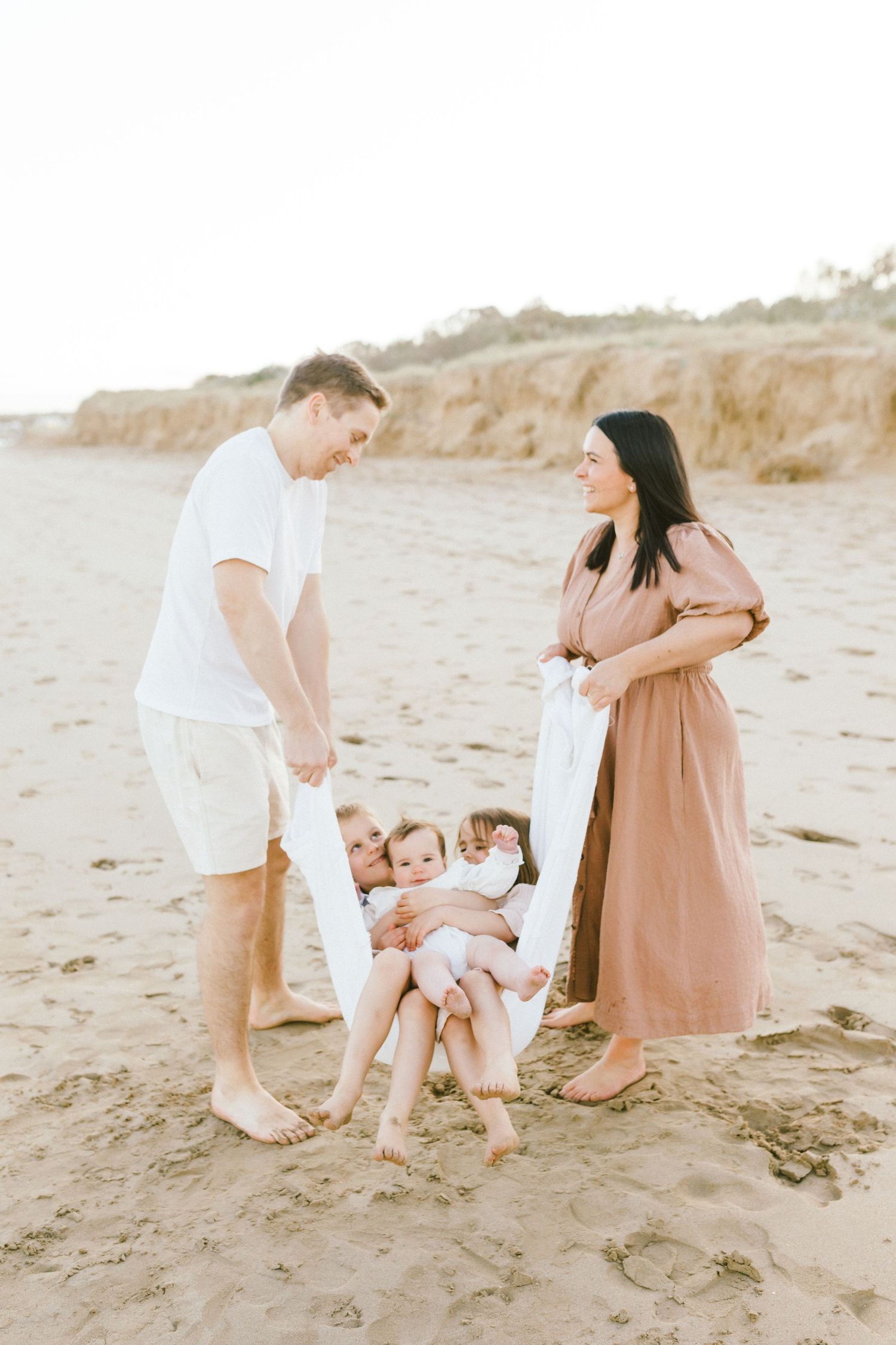 A family in white and tan outfits plays together on a sandy beach at sunset, creating tender moments.