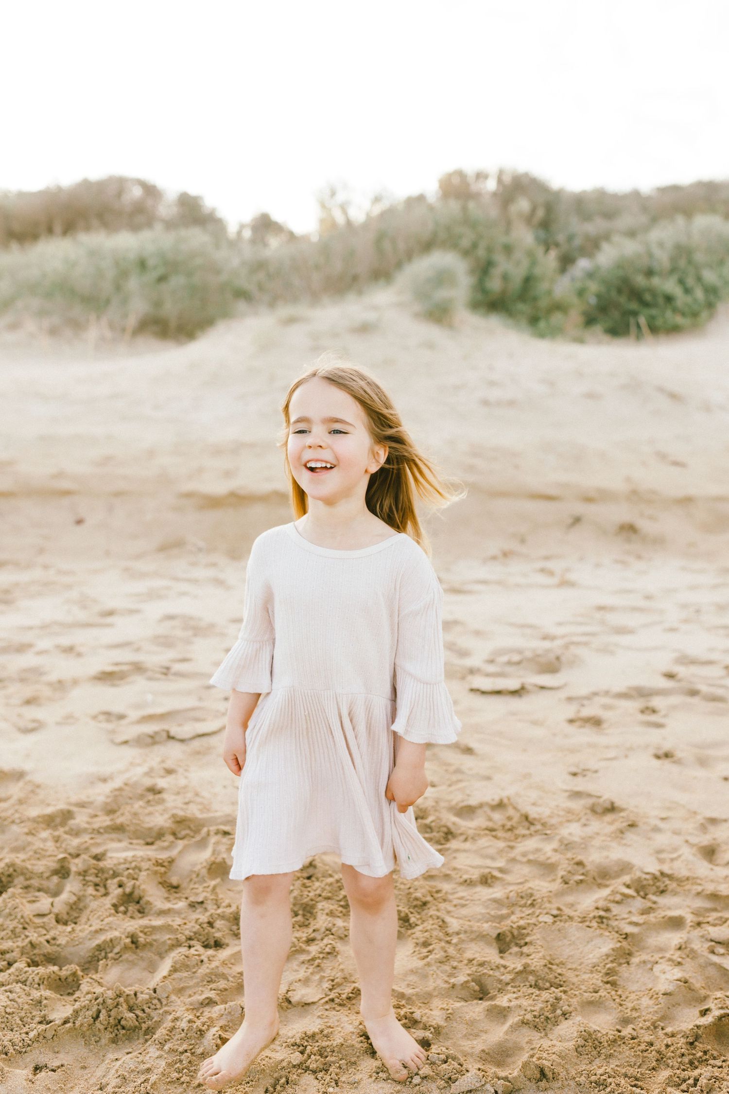 A child in a white dress plays joyfully on a sandy beach during a sunny day.
