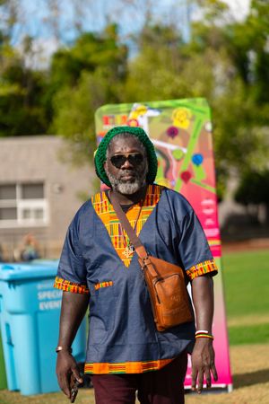 Festival worker wearing orange safety vest and green hat stands outdoors near colorful event setup.