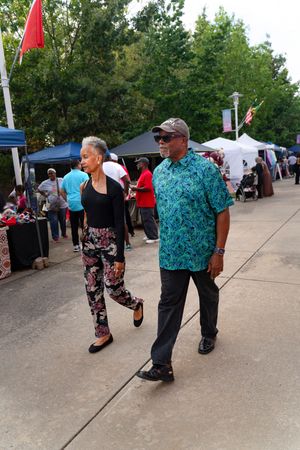 People walking at an outdoor festival with vendor tents and trees in the background.
