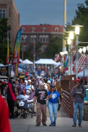 Crowd of people walking along a festival street with white tents and colorful flags in the background.