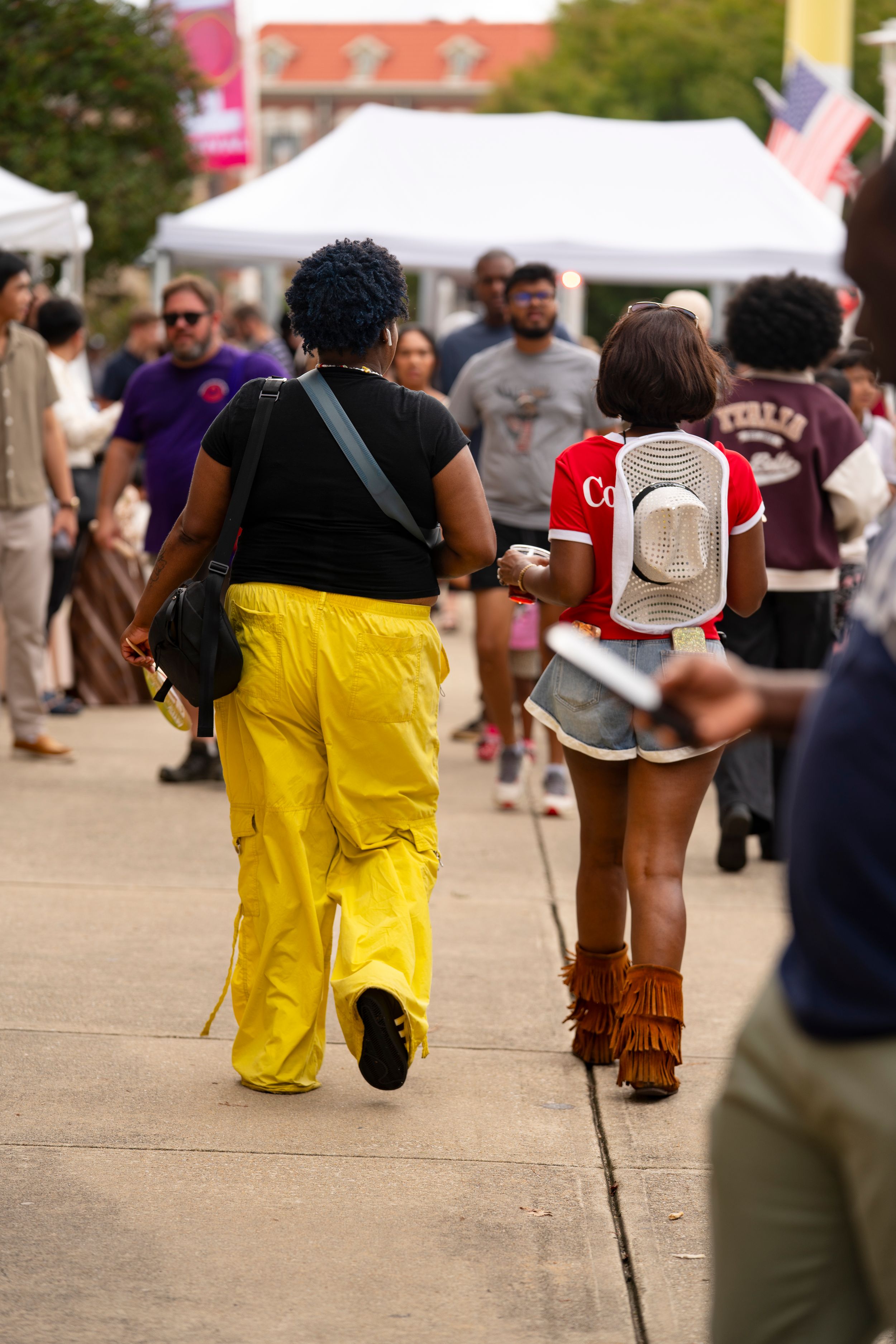 Two people walking at outdoor festival, one wearing bright yellow pants.