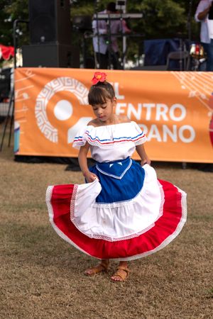 Dancer in traditional red, white and blue dress performs in front of orange banner.