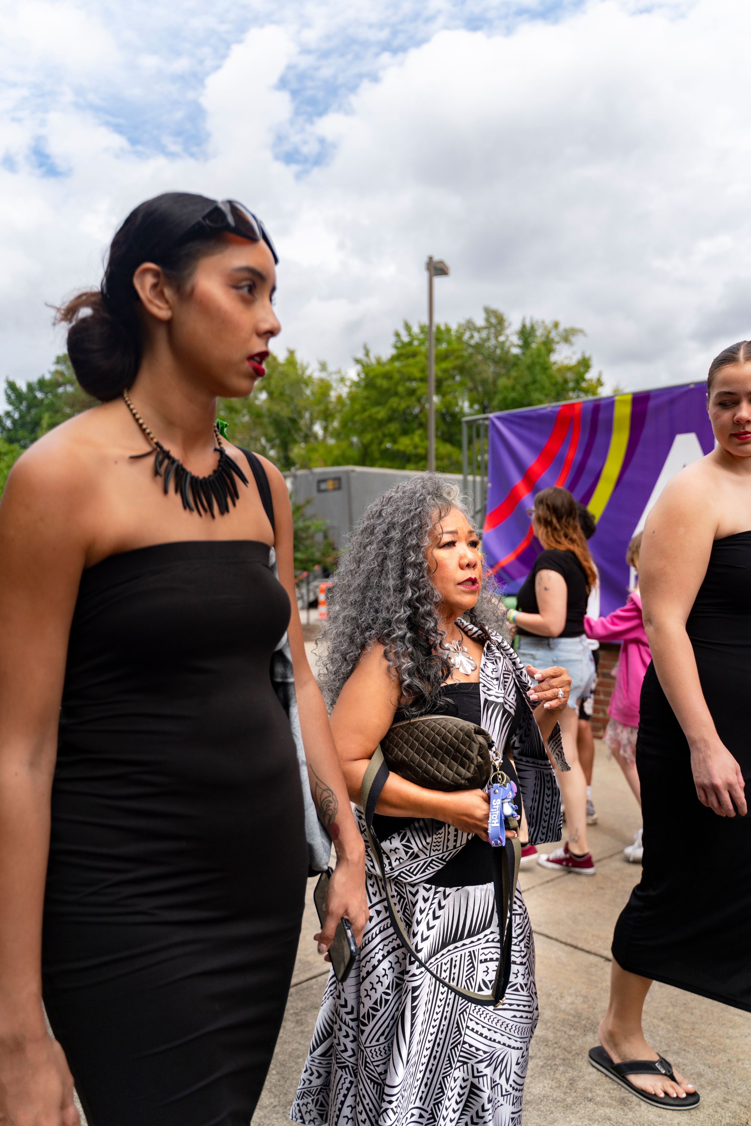 Group of people in formal attire standing together at an outdoor event with a colorful flag visible in background.