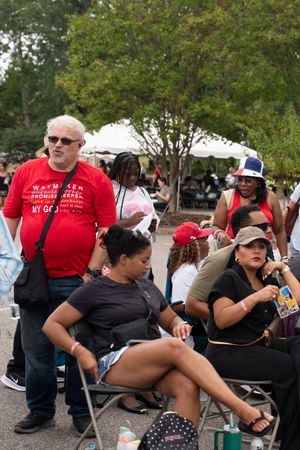 Festival attendees sit in camping chairs while waiting in line at an outdoor event on a sunny day.