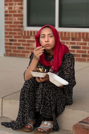Person in patterned dress with red hair eating from plate while sitting on concrete steps.