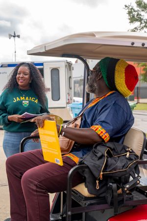 People in casual attire gather around a golf cart at an outdoor event wearing Jamaican-themed accessories.