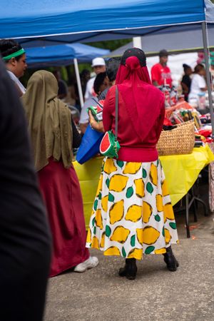 People in colorful clothing browse vendors at an outdoor market under blue tents, with a vibrant lemon-print skirt visible.