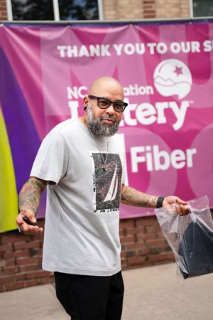 A person in a gray t-shirt and sunglasses gestures in front of a pink promotional banner display.