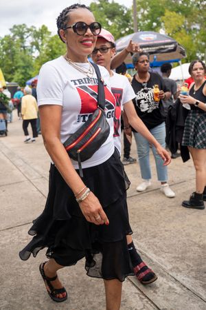 A person in sunglasses and a graphic t-shirt smiles while walking at an outdoor event with others in the background.