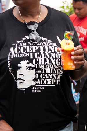 A black t-shirt with an Angela Davis quote and portrait design, shown with someone holding an orange tropical drink.