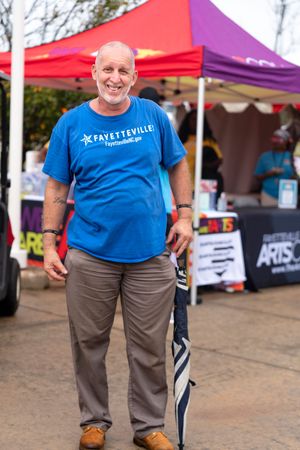Person wearing a blue Fayetteville t-shirt and brown pants stands at an outdoor event with vendor tents in background.