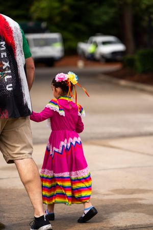 A person wearing a vibrant pink traditional Mexican dress with colorful rainbow stripes walks along a path.