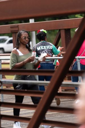 People gathered on a wooden deck or boardwalk viewed through brown railing slats on a sunny day.