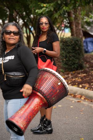 Musicians perform with red drums at an outdoor event wearing black outfits and sunglasses.