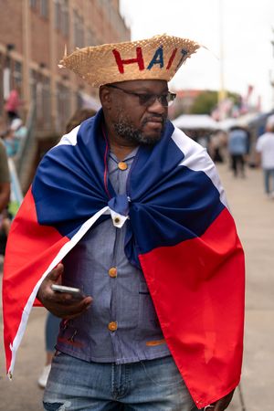 Person wearing a straw hat with 'HAI' text draped in Haiti's red and blue flag colors on a city street.
