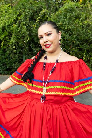 A vibrant red traditional Mexican dress with colorful ribbon trim and ruffled details is displayed against green foliage.
