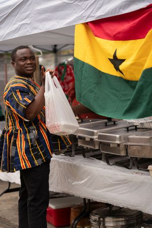 Food vendor in traditional African print shirt serving at outdoor event with Ghana flag displayed.
