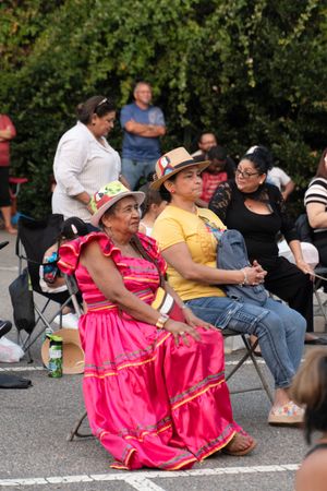 People sitting in folding chairs at an outdoor cultural event wearing bright pink and yellow traditional dresses.