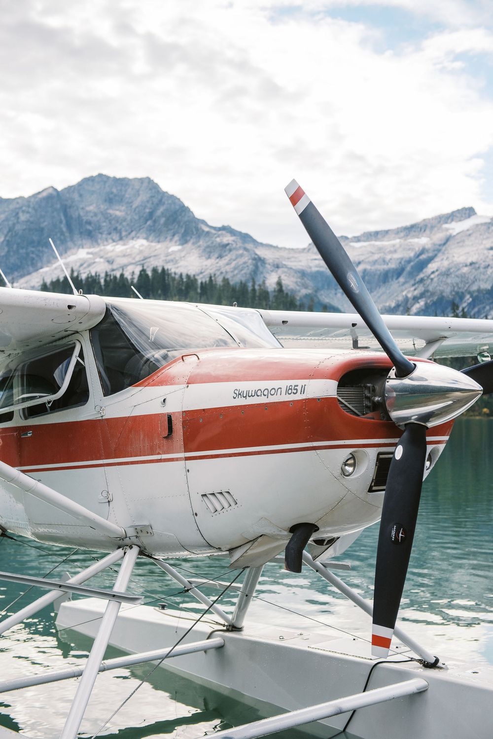 Seaplane with red and white paint docked on a mountain lake with snow-capped peaks in the background.