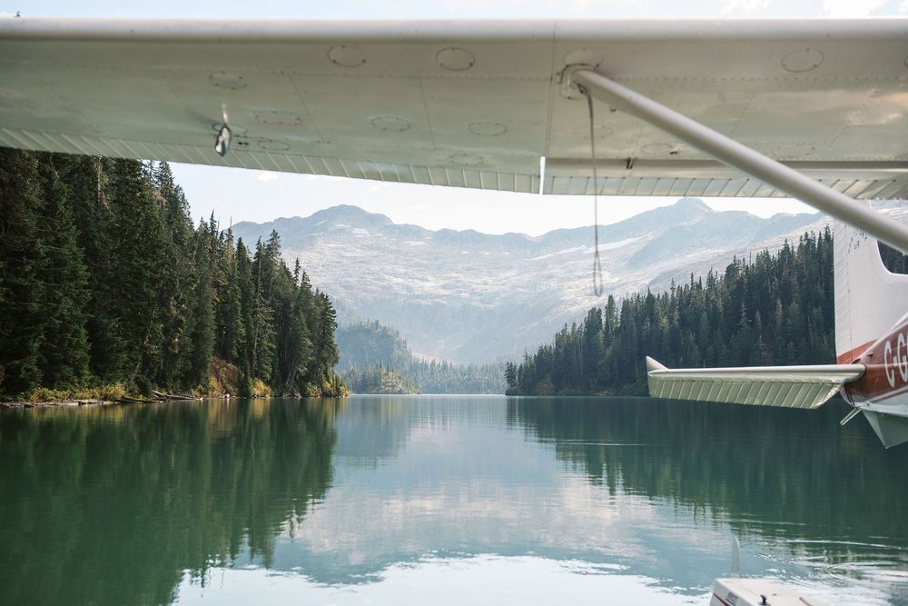 Scenic mountain lake surrounded by evergreen forests reflecting in calm waters with seaplane floats visible.