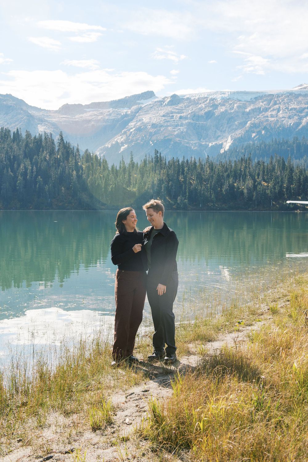 A couple shares a romantic moment by a serene mountain lake surrounded by evergreen forests and snow-capped peaks.