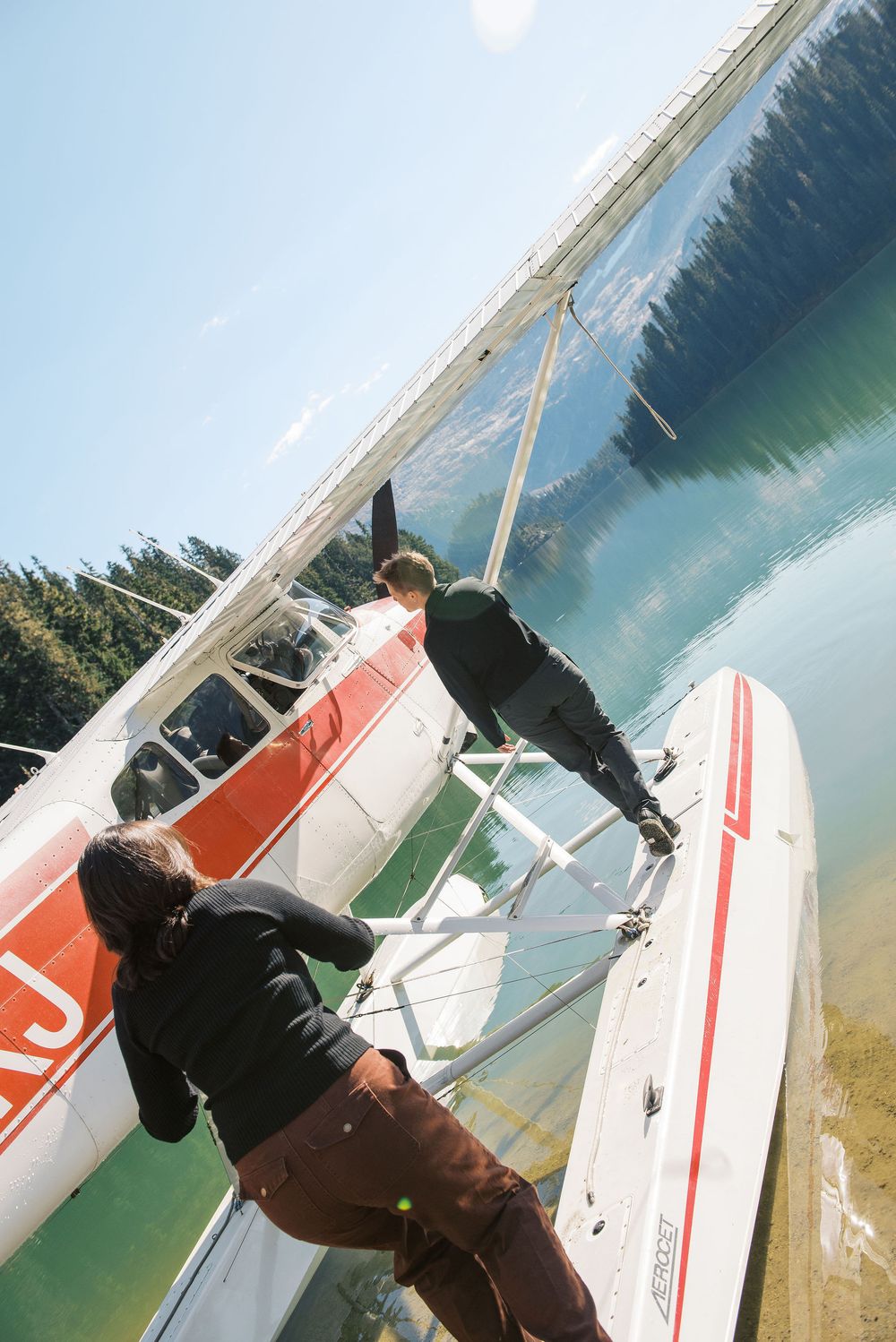 A seaplane floats on calm turquoise waters with mountains and forest visible in the distance.