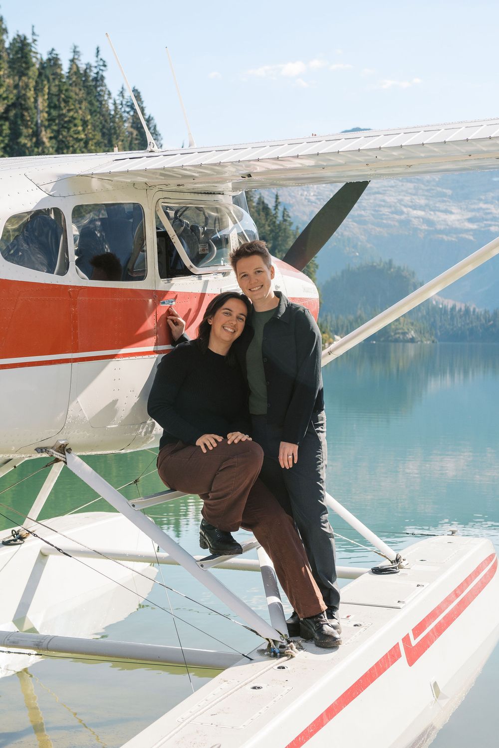 A couple poses on the floats of a red and white seaplane against a backdrop of mountains and lake.