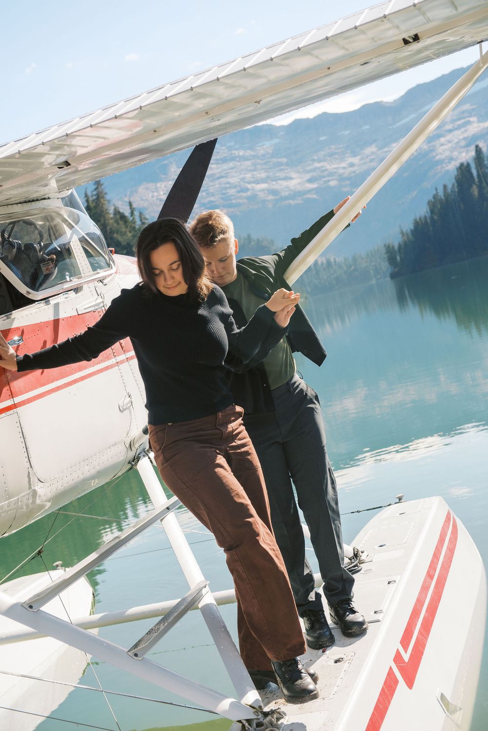 Two people stand on a seaplane float dock beside turquoise mountain lake waters on a sunny day.