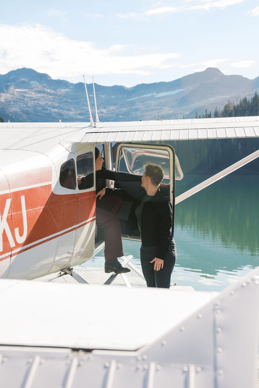 Two people stand near the door of a red and white Cessna seaplane overlooking a mountain lake.