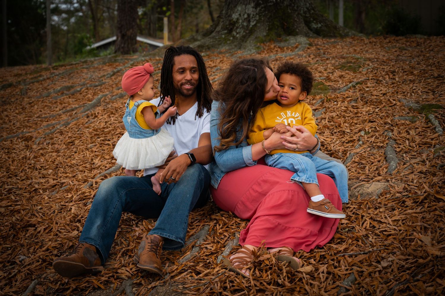 Family relaxes together on ground covered with autumn leaves during outdoor photo session.