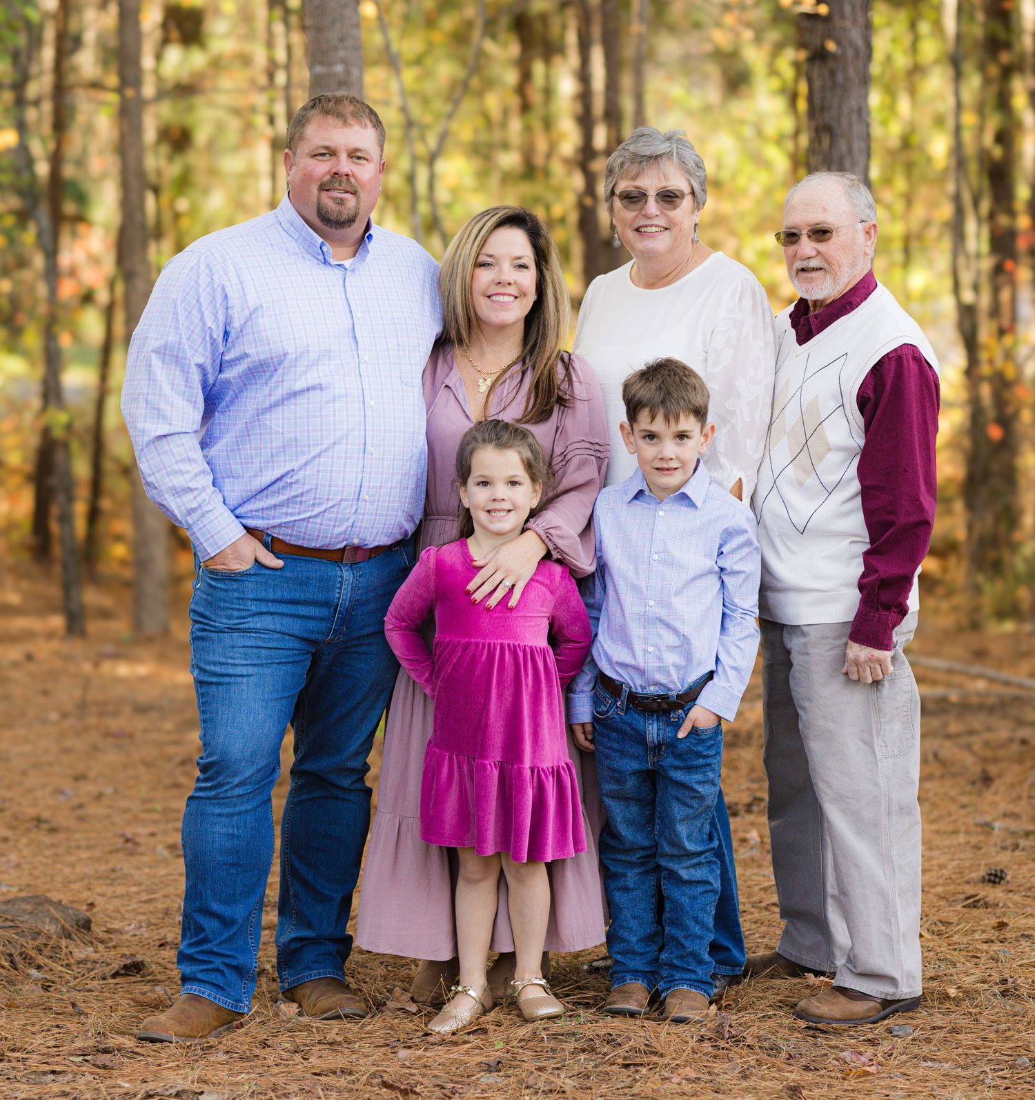 Multi-generational family portrait in autumn forest setting with pine trees in background.