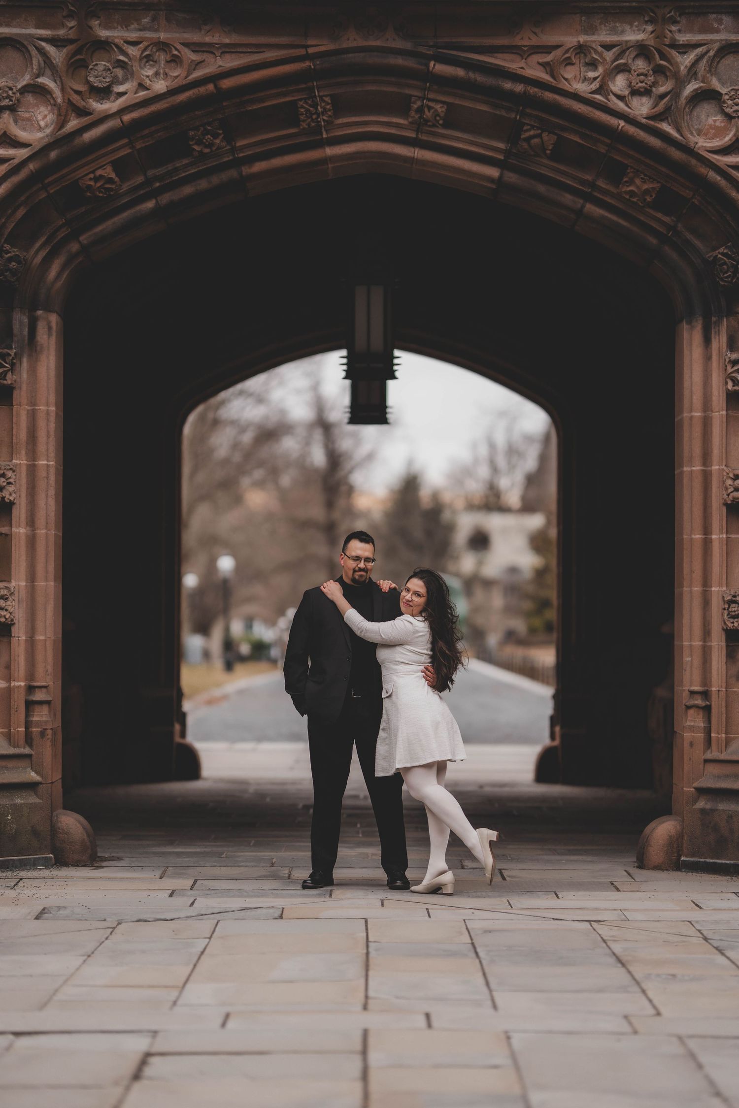 A couple stands together under an ornate gothic stone archway during a romantic photoshoot on a cloudy day.