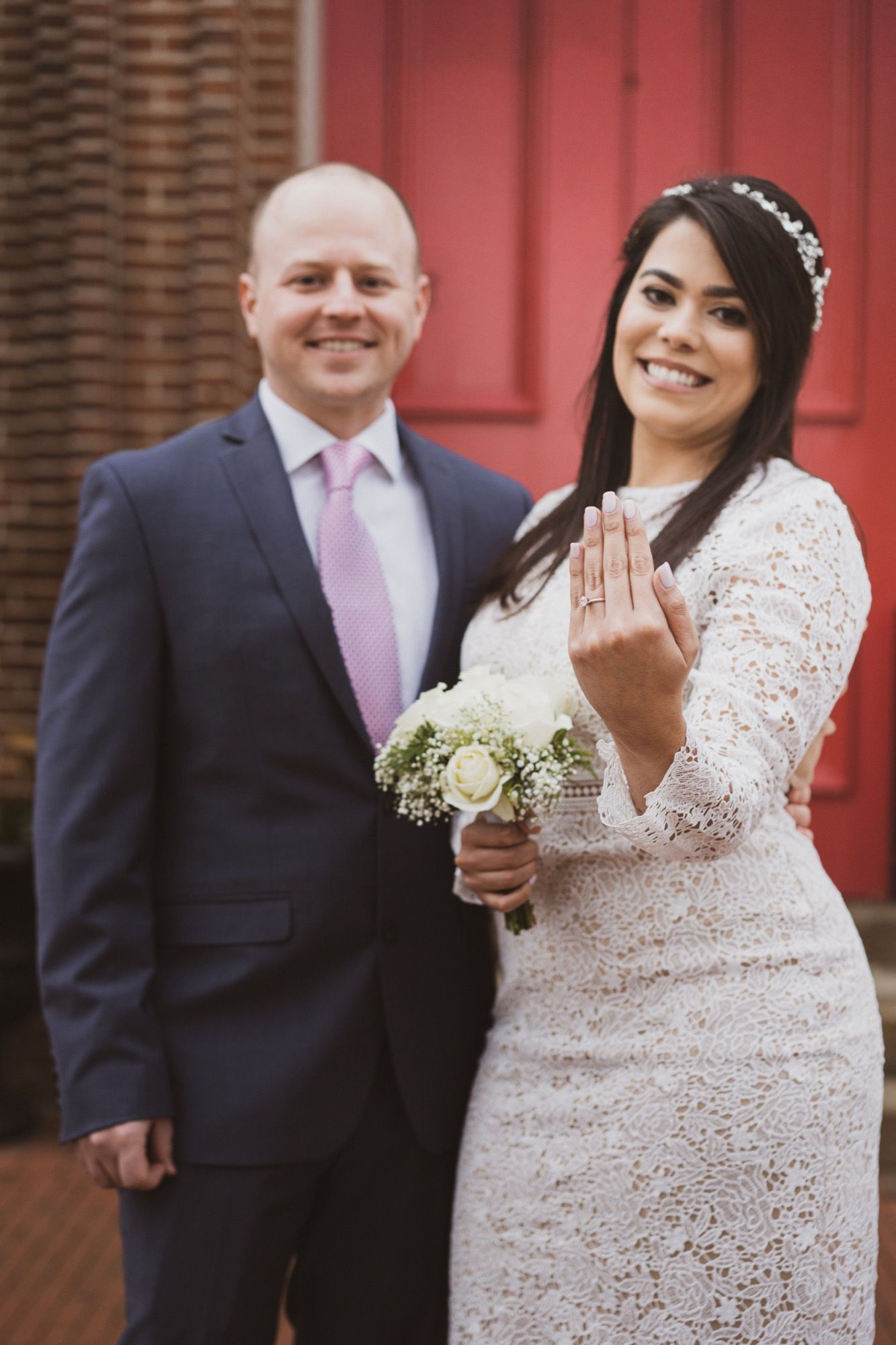A newlywed couple poses with bride showing off ring while holding white bouquet against red brick background.