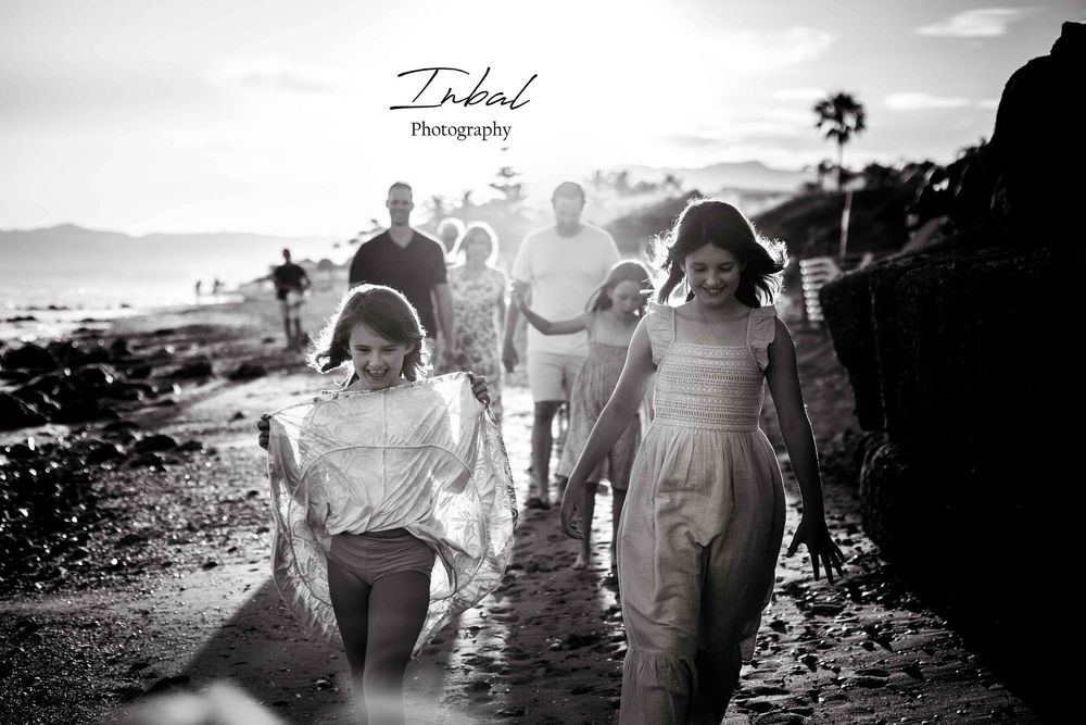 Black-and-white photo of a family walking along the beach at sunset, one behind the other in an unposed, natural way, with the sun setting b