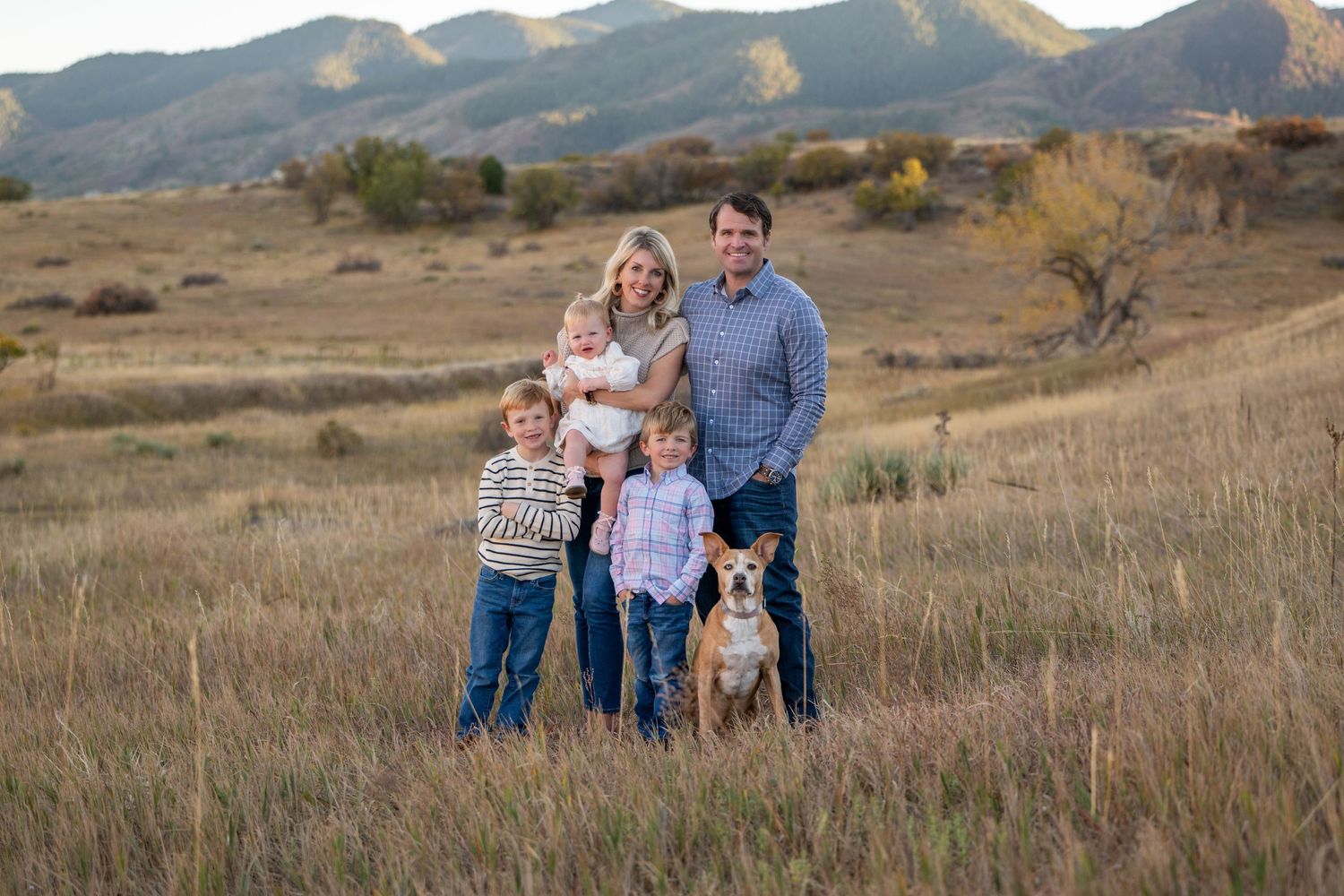 A family and their dog stand together in a grassy field with mountains in the background during golden hour.