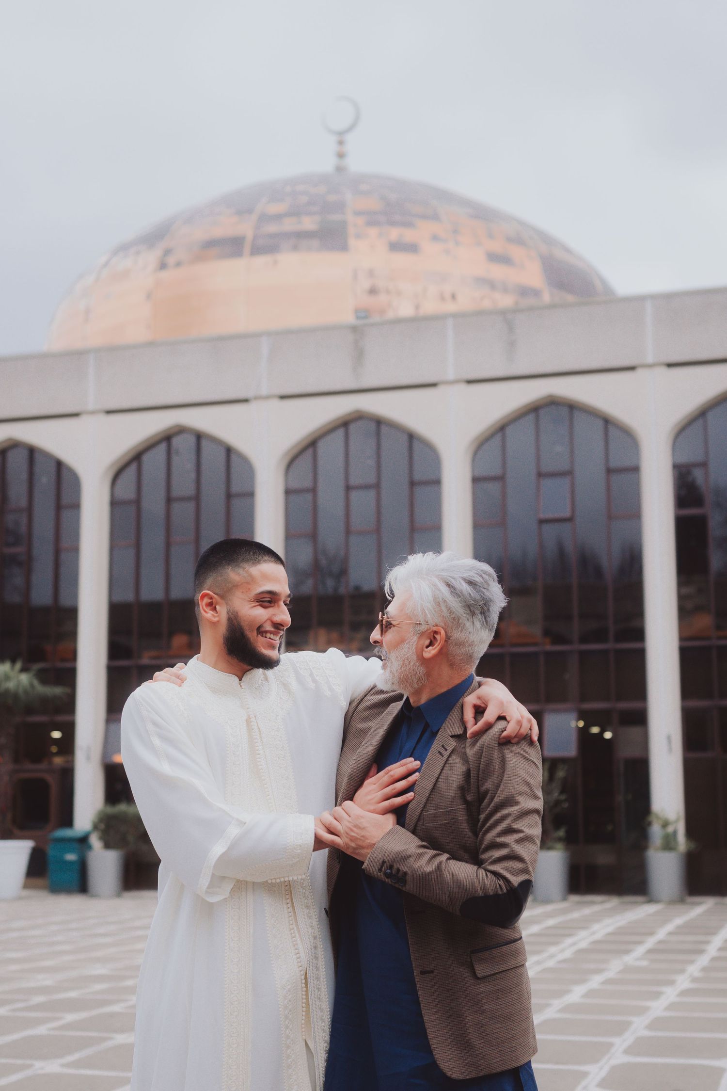 Two people share a warm moment outside a mosque with distinctive domed architecture.
