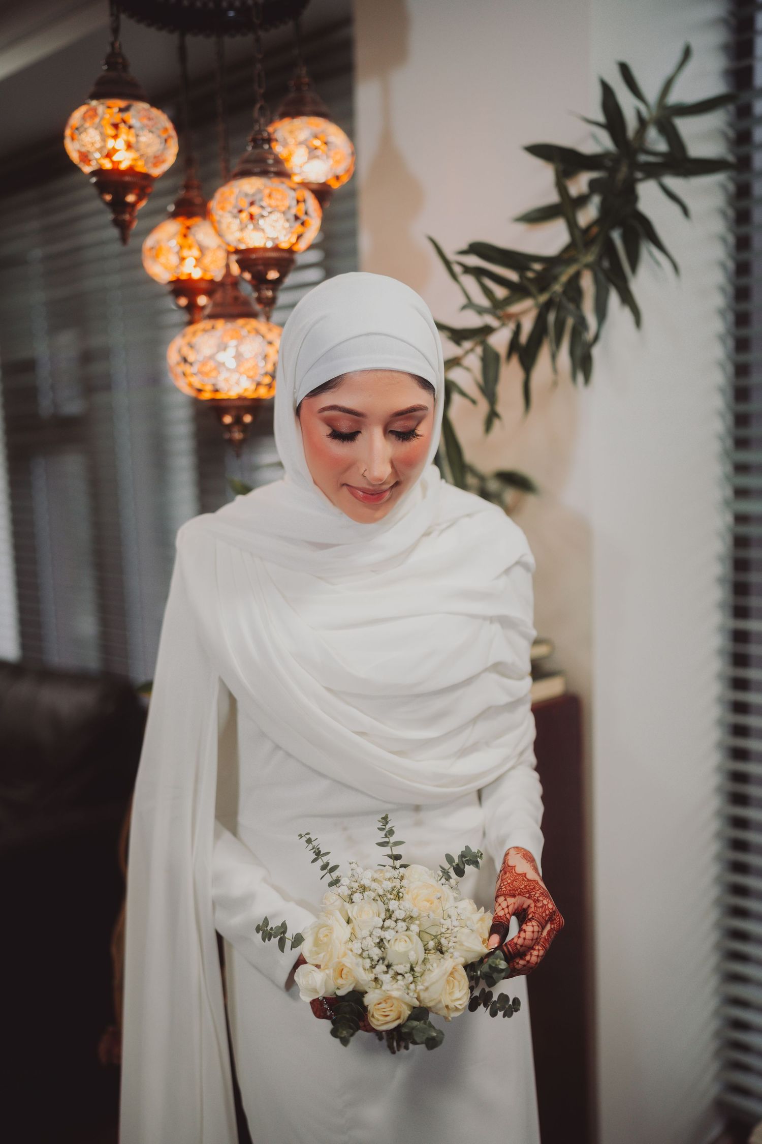 Bride in elegant white hijab and dress holds bouquet beneath Moroccan lanterns.