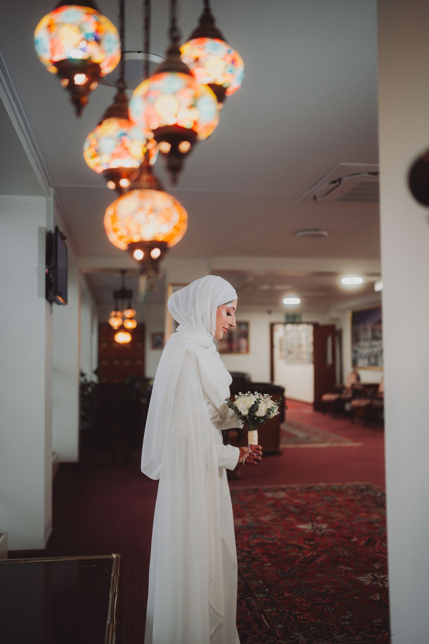 Figure in flowing white dress and hijab stands in hallway under glowing Moroccan lanterns.
