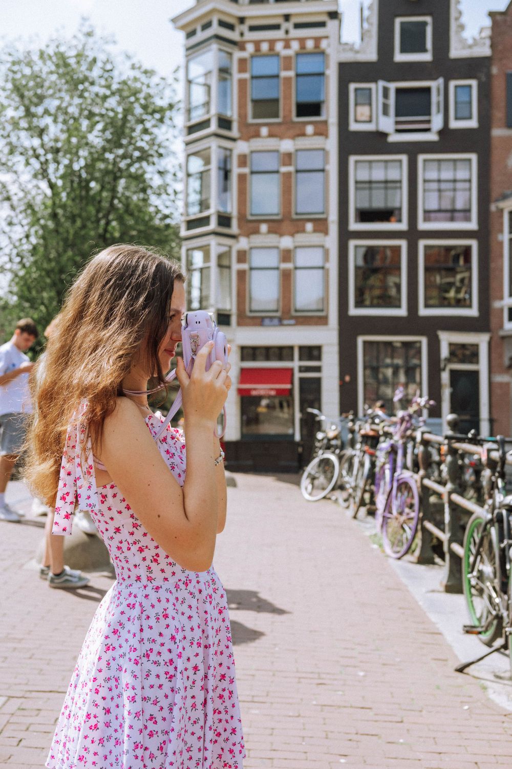 Woman in floral summer dress walking along Amsterdam canal with historic buildings and bicycles.