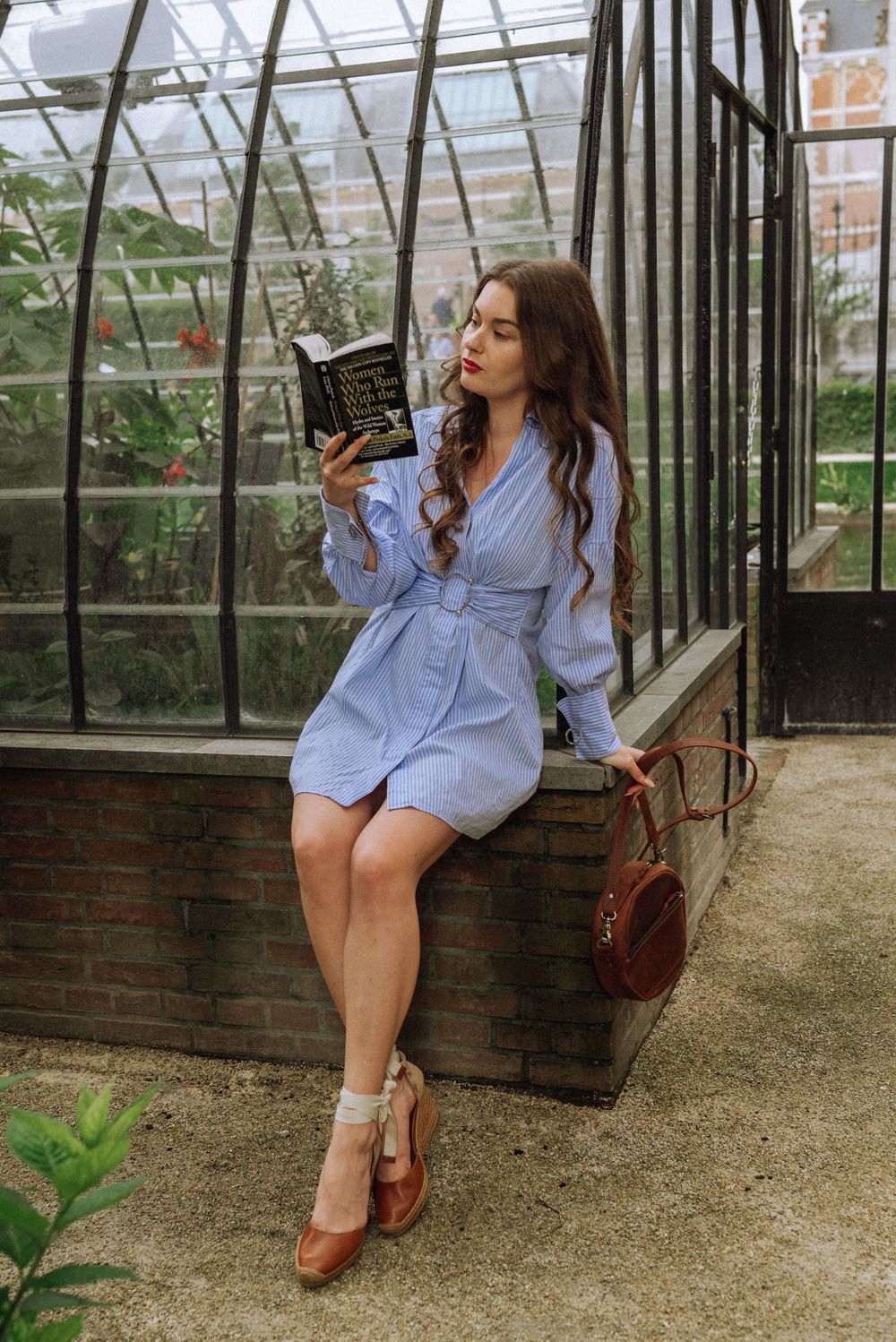 Fashion model poses in blue shirt dress near vintage greenhouse with brown leather accessories.